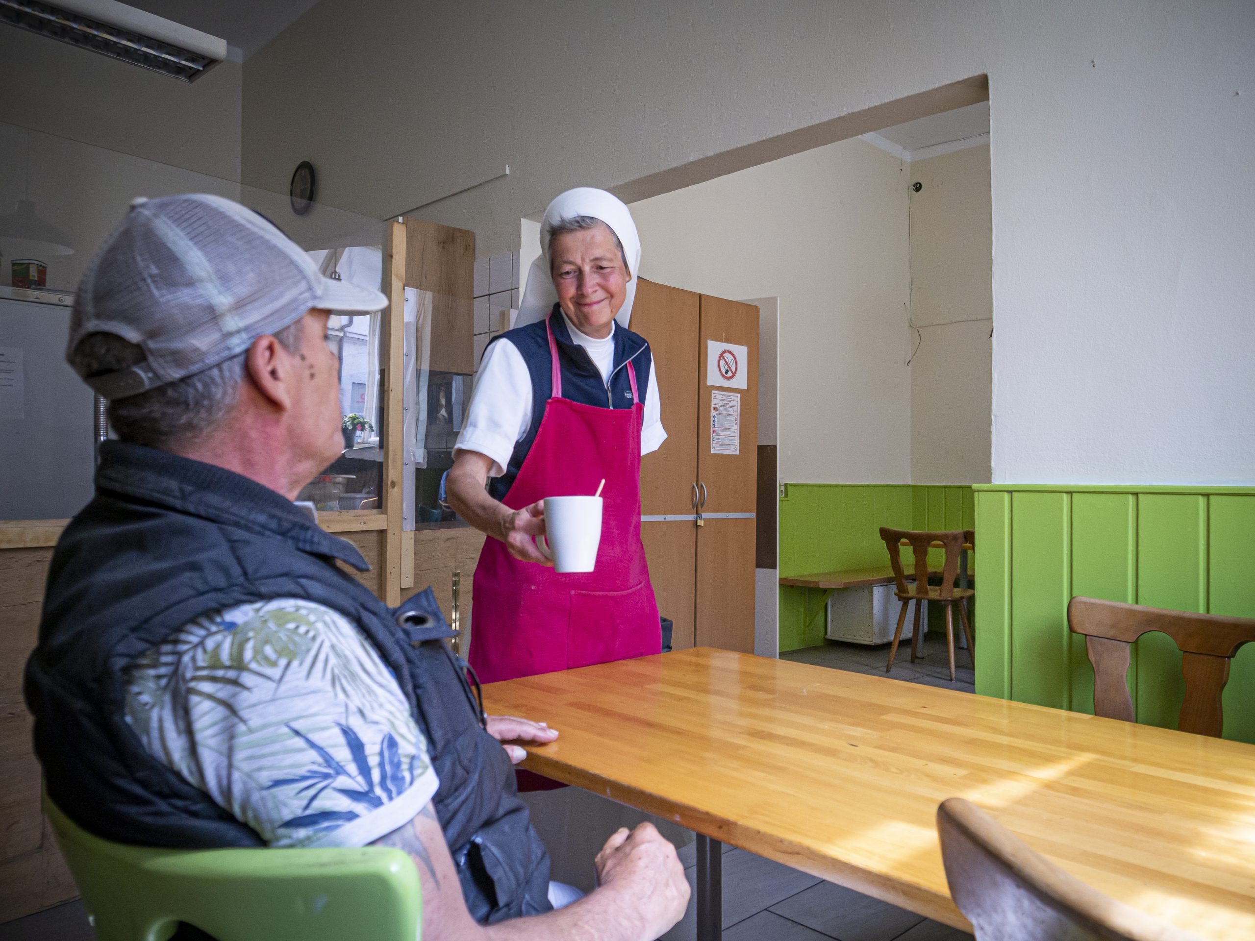 Ältere Dame mit Kochschürze und Kopfbedeckung reicht älterem Herrn in Cafeteria einen Tasse an seinen Platz